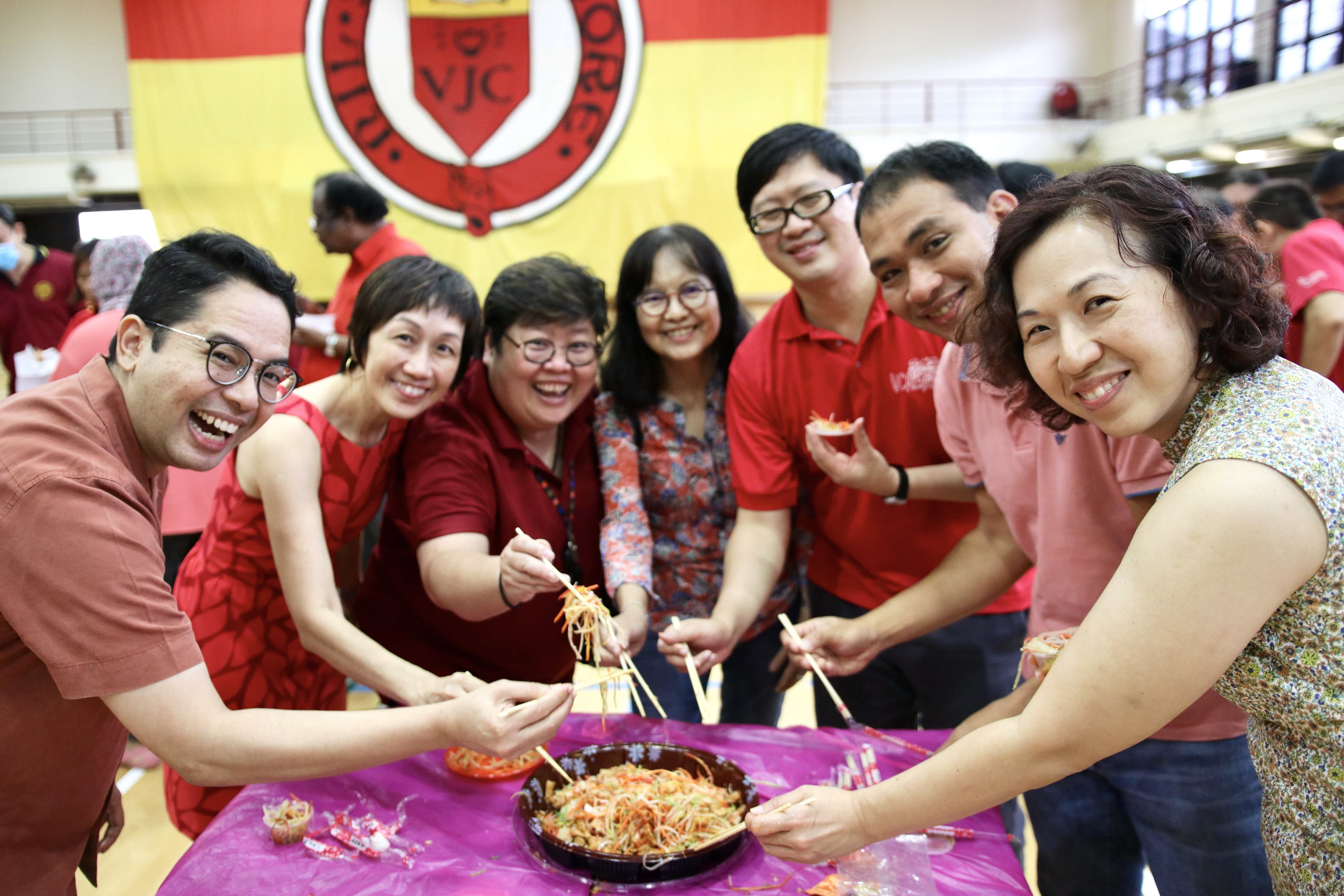 Six smiling people use chopsticks to toss yusheng salad at a table during a Chinese New Year celebration.